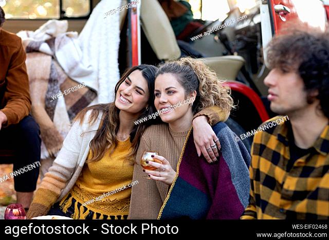Smiling woman sitting with arm around friend on picnic outside campervan