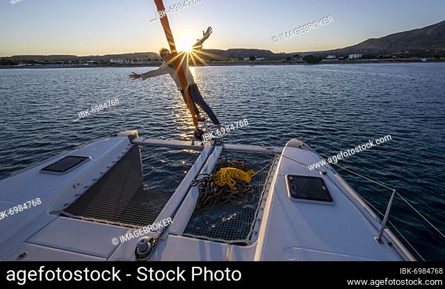 Young man standing by the foresail, sunset with sun star, Sailing a sailing catamaran, Dodecanese, Greece, Europe