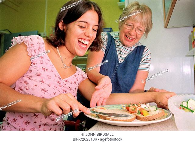 Carer assisting teenage girl with physical disability to prepare a sandwich in kitchen of residential respite care home