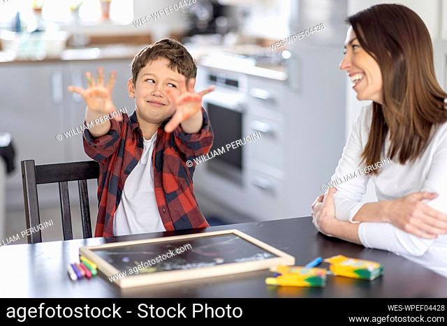 Boy showing dirty hands while looking with sideways glance at mother in kitchen