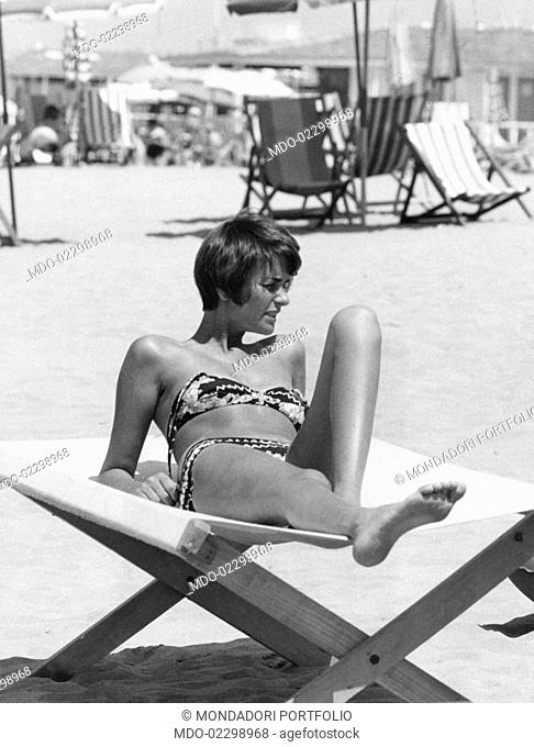 A woman wearing a bikini looks absent-midendly elsewhere, while sunbatheing on the beach, lying on a beach chair; behind her