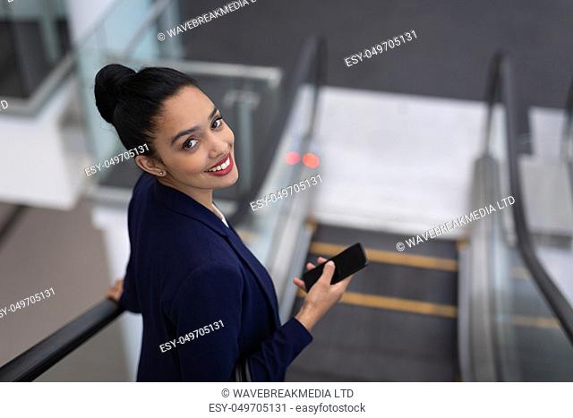 Portrait of happy pretty young mixed-race businesswoman with mobile phone looking at camera on escalator in modern office. She is smiling at camera