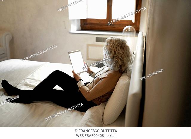 Mature businesswoman sitting on bed looking at digital tablet