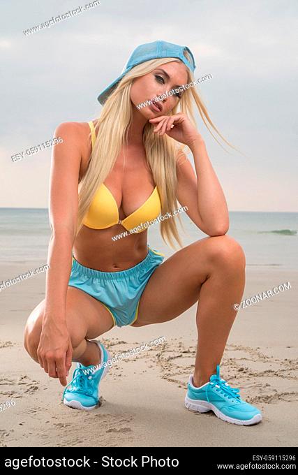 Sexy blond woman wearing yellow bra, sporty blue shorts and cap looking into the camera while crouching down on the sandy beach over cloudy sky and sea...