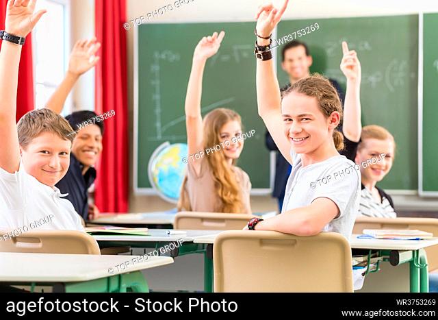 School class teacher giving lesson in front of a blackboard or board teaching students or pupils, they are raising their hands as they know all the answers