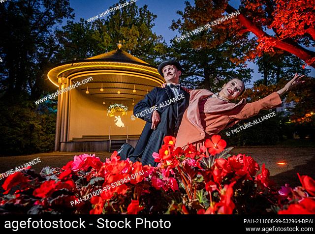 08 October 2021, Saxony, Bad Elster: Actors dressed as Charlie Chaplin and Anna May Wong pose in front of the illuminated bandstand at the Royal Spa House in...