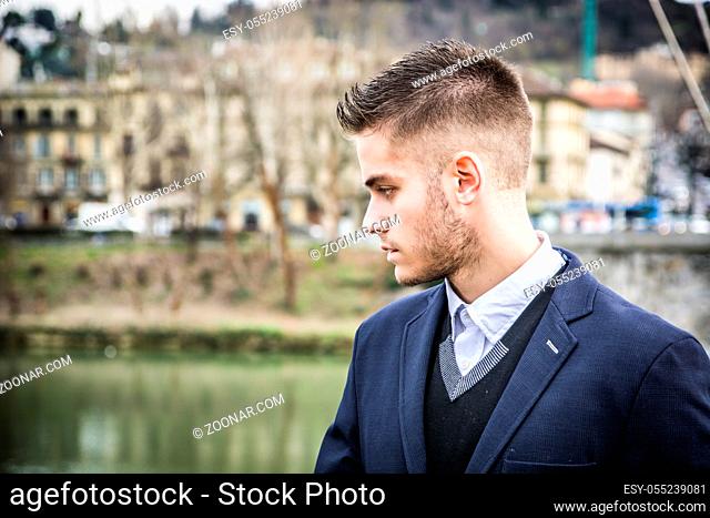 Three-quarter length of contemplative light brown haired young man wearing grey jacket and denim jeans standing beside picturesque river in Turin, Italy