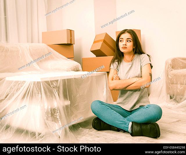 Brunette young woman trying to be patient while relocating to new apartment. Female sitting on the floor in new flat with lots of unpacked boxes in the room...