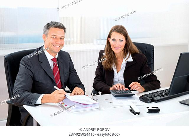 Two Businesspeople Calculating Tax Together At Desk In Office