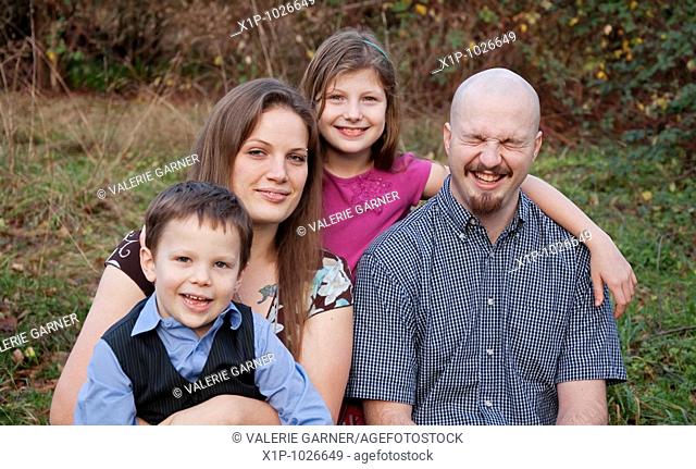 A young Caucasian family of 4 portrait outdoors, dad is laughing.  Two kids, boy and girl with mom and dad