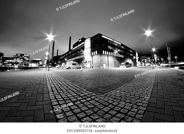 Dramatic low angle black and white night photo of. Large building between lamp posts and stone pavement on foreground