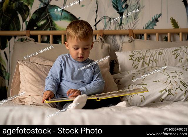 Cute boy reading book sitting on bed at home