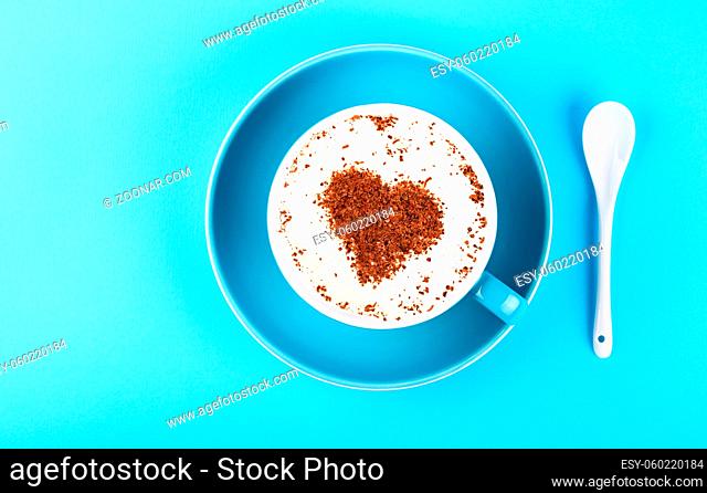 Close up one full cup of latte cappuccino frothy coffee with heart shaped chocolate and saucer with white spoon over pastel blue paper background
