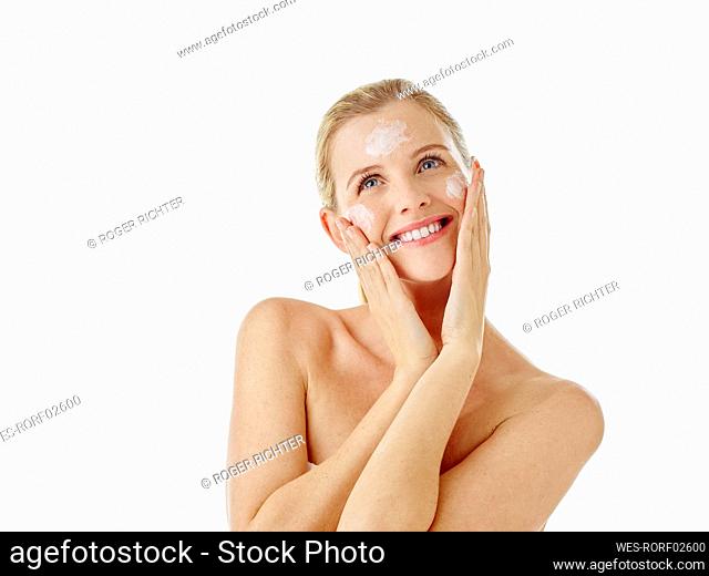 Young woman smiling while applying face cream standing against white background