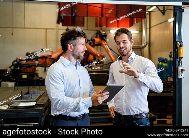 Businessmen with digital tablet discussing while standing against robotic arm machine at factory