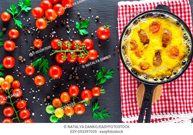 fried omelette with sausages and chicken eggs in a round cast-iron pot and ripe red cherry tomatoes on a black background, top view