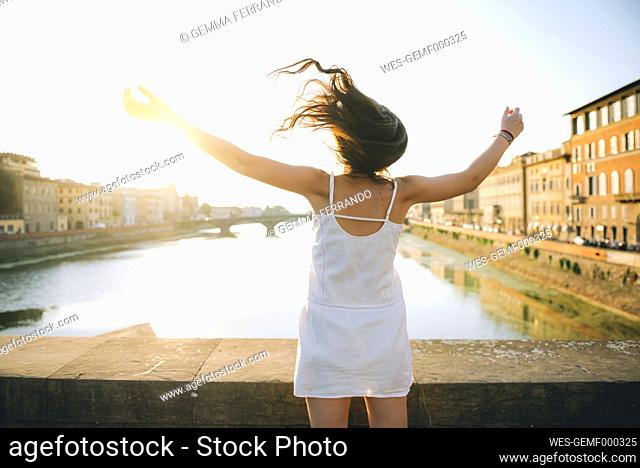 Italy, Florence, back view of happy woman wearing white summer dress standing on a bridge at sunset