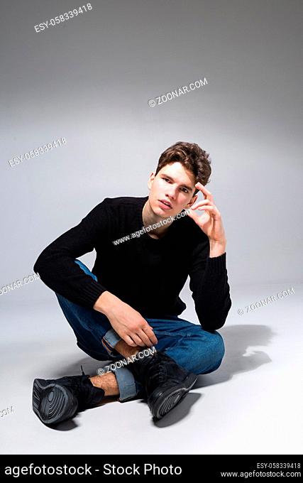 Stylish guy posing in studio sitting on the floor in denim pants and black sweatshirt. Male student portrait full on white background
