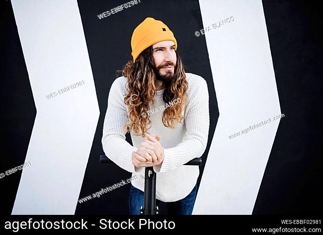 Hipster man in knit hat looking away while leaning on electric scooter