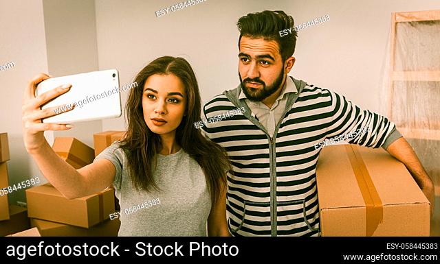 Relocation young positive couple making selfie shot in new home. Man and woman in the room full of unpacked boxes, man holding box and woman holding phone...