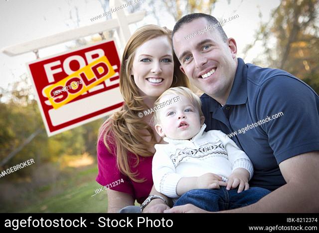 Happy young caucasian family in front of sold real estate sign