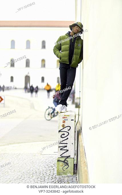 young stylish man standing on electricity box in city, tired, nonconformity, leaning against wall with closed eyes, in Munich, Germany
