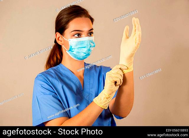 Brunette nurse in blue shirt putting on rubber gloves. Caucasian girl touching her hand with fingers on light background