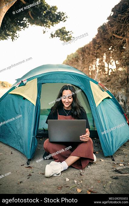 Smiling young woman working on laptop while sitting in tent during weekend