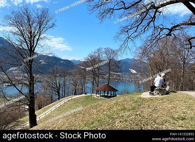 Couple take a rest on a bench. Hikers on the Hoehenweg over the Tegernsee with a view of Rottach Egern on April 1st, 2021