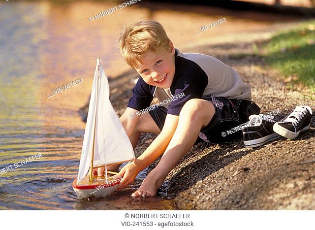 park-scene, portrait, full-figure, 10-year-old blond boy wearing blue-beige shirt and scater jeans plays bare-feeted with his red toy sailing boat at the water...