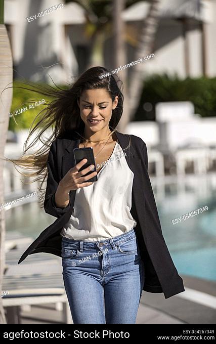 Trendy brunette with flying hair walking in hotel poolside and browsing smartphone in sunlight
