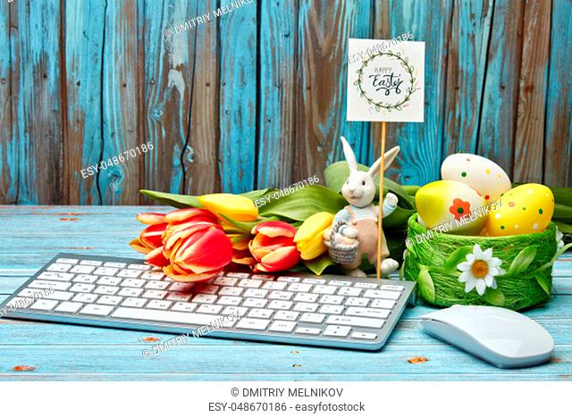Colored easter eggs, easter rabbit, spring flowers, computer keyboard and mouse on a wooden background with copy space. Greeting card, Easter background