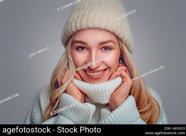 Attractive blond woman in a warm woollen polo neck sweater and knitted cap standing in front of a grey background snuggling into the collar with a wide happy...