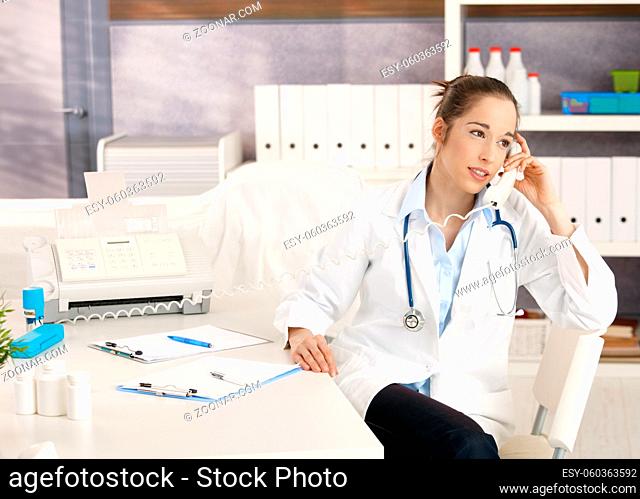 Young female doctor sitting at desk in doctor's room calling, looking away, smiling