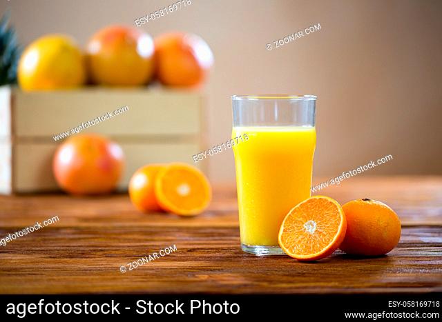 Orange cut in half lying on a wooden table next to glass of freshly squeezed juice. Yellow liquid from fruit with brown box in background