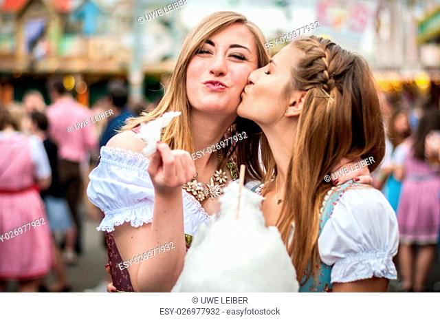 Two young women in traditional Bavarian clothes, dirndl or tracht, kissing with cotton candy floss at the Oktoberfest