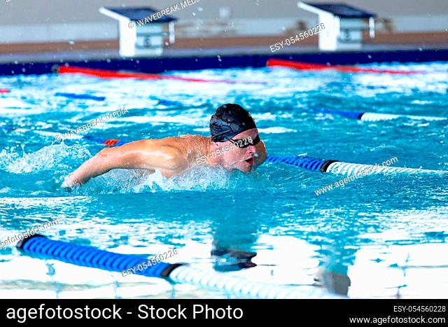 Side view of a Caucasian male swimmer at swimming pool, wearing a black swimming cap and goggles, splashing, swimming butterfly
