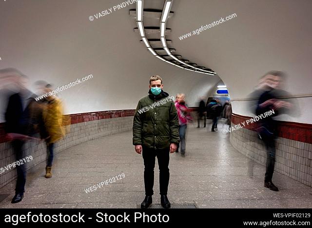 Young man with face mask standing isolated in subway underpass, with people moving around him