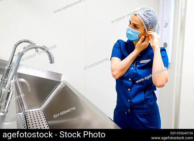 Female surgeon puts on a mask near the washbasin, preparing for surgery operation. Doctor in uniform, medical clinic worker, medicine and health