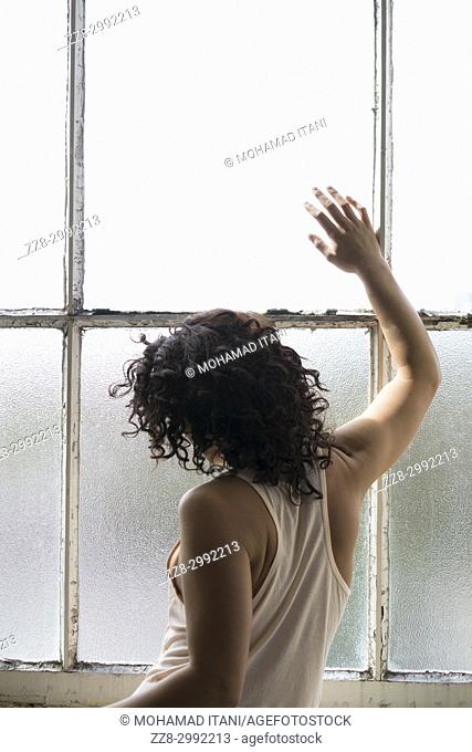 Rear view of a young woman standing by the window hand touching the glass