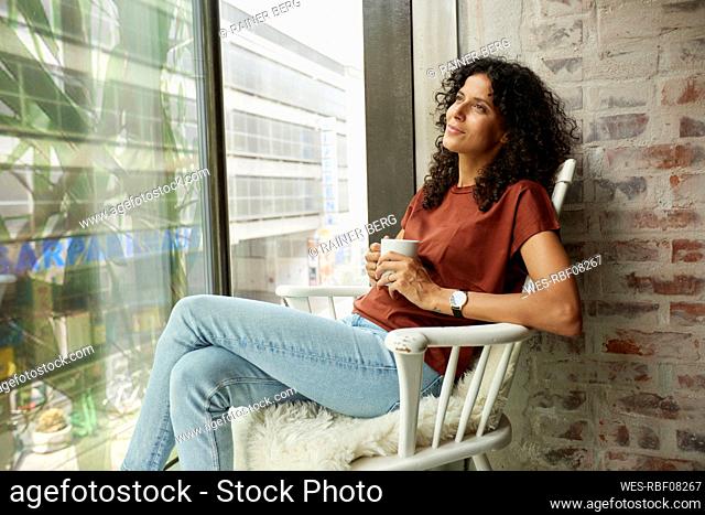 Thoughtful female professional with coffee cup sitting on chair by window