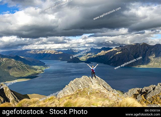 Hiker stands on a rock and stretches her arms in the air, view over Lake Hawea, lake and mountain landscape in the evening light, view from Isthmus Peak, Wanaka
