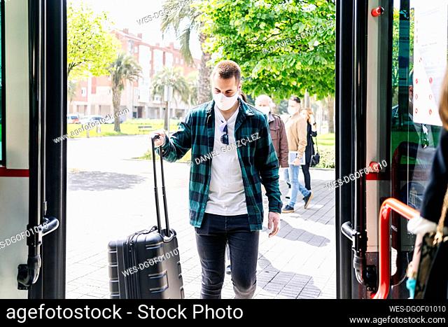 Young man wearing protective mask getting into bus with trolley bag, Spain