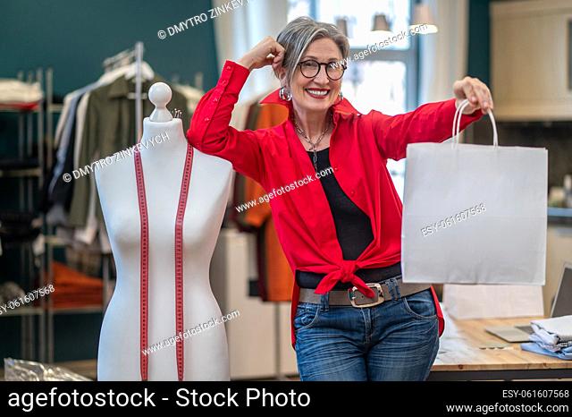 Determination. Gray-haired elegant woman smiling showing package at camera standing leaning on mannequin in atelier