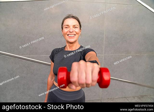 Happy woman showing red dumbbell in front of wall