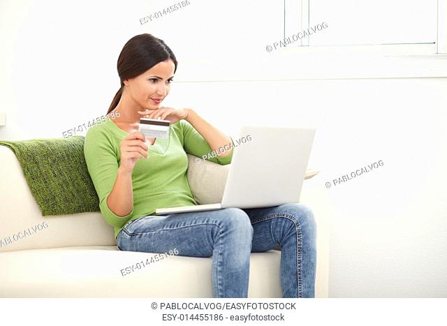 Young woman in green shirt holding a laptop and a credit card while sitting indoors