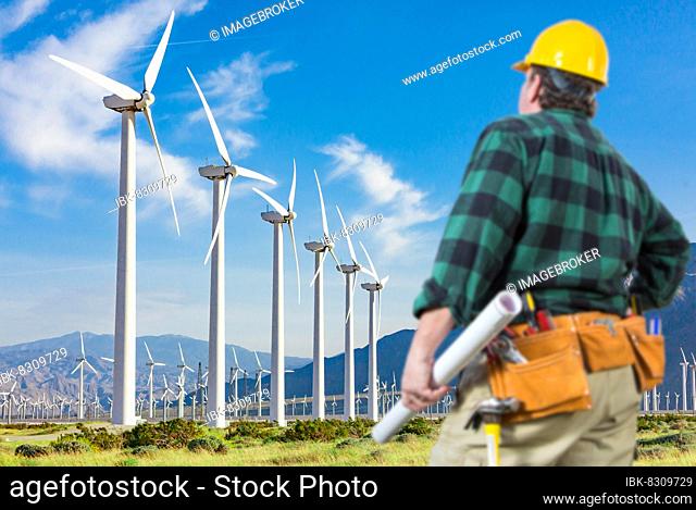 Male contractor wearing tool belt and hard hat facing alternative energy wind turbines