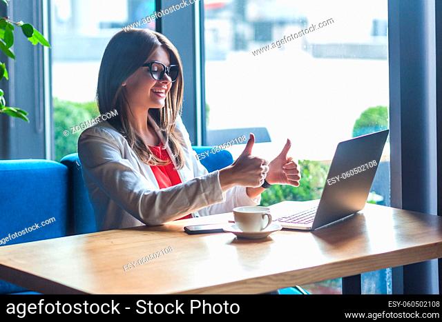 Like it. Portrait of happy satisfied beautiful stylish brunette young woman in glasses sitting, looking at her laptop screen, toothy smile and thumbs up