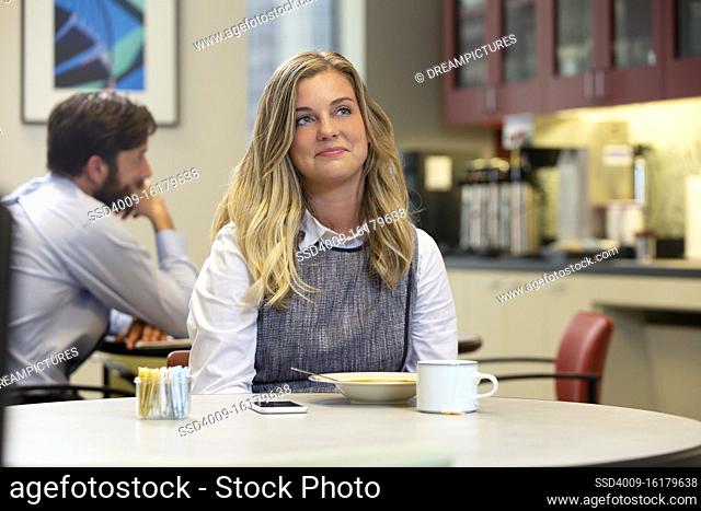 Young Caucasian woman looking off camera with a smirk, while sitting in break area of office with lunch of soup and Hot Tea