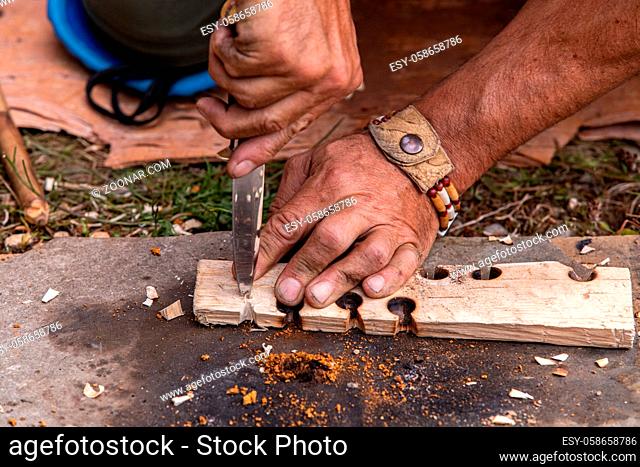 Man hand holding knife while giving demonstration to light fire on wooden slab with burnt holes using native traditional technique at world festival